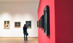 Person standing in an art gallery viewing framed paintings on a white wall, with a red accent wall in the foreground.