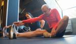 2 people stretching side by side on a gym floor with weights and a window in the background.