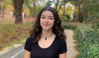 A smiling young woman with curly brown hair standing outdoors on a college campus pathway, surrounded by green foliage and autumn-colored trees. She is wearing a black top and a simple necklace, exuding a friendly and approachable demeanor.