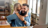 A young daughter kissing her bearded father on the cheek as he smiles, with a laptop visible on the desk in a cozy home office setting.