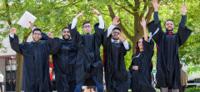 Group of six graduates wearing black caps and gowns outdoors, joyfully jumping and raising their hands in celebration under green leafy trees on a sunny day.