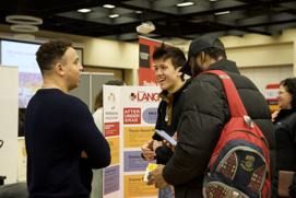 Students engaging in a conversation at a professional programs fair, discussing options like MBA, graduate, and thesis-based programs with informational displays in the background.