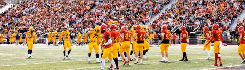 University of Guelph football players in yellow and blue uniforms face off against red team members on the field, with a cheering crowd in the background during a homecoming game.