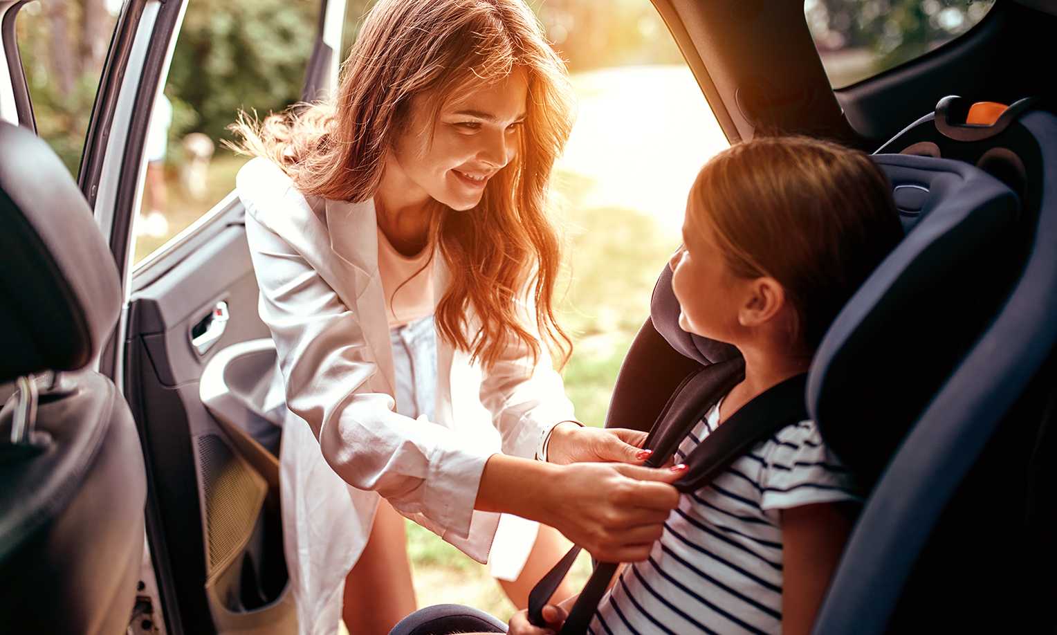 Mother buckling her young child into a car seat with a warm smile on her face.