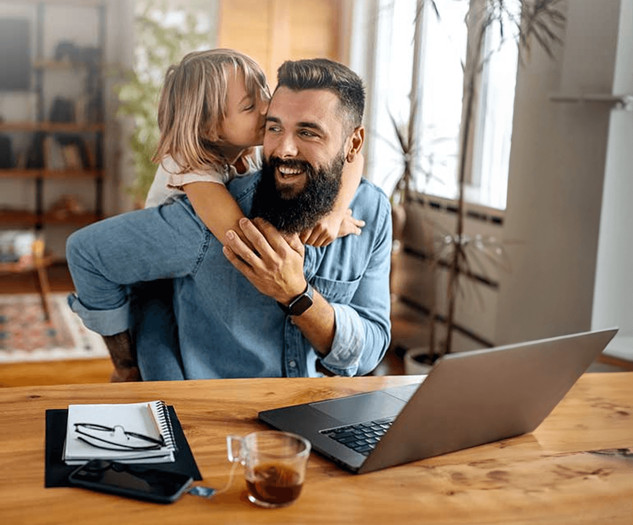 Smiling man sitting at a table, hugged and kissed on the cheek by a young girl, with a laptop, notebook, and cup of tea in front of him.