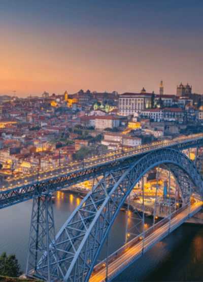 A scenic view of Porto, Portugal at twilight, showcasing the iconic Dom Luis l Bridge spanning the Douro River. The city is illuminated with golden lights, highlighting the historic architecture and steep hillsides, creating a vibrant contrast against the fading blue sky.