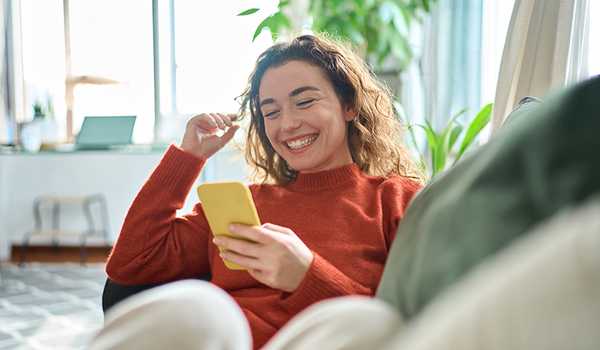 Woman in an orange sweater smiling while using her phone, sitting comfortably on a couch at home.