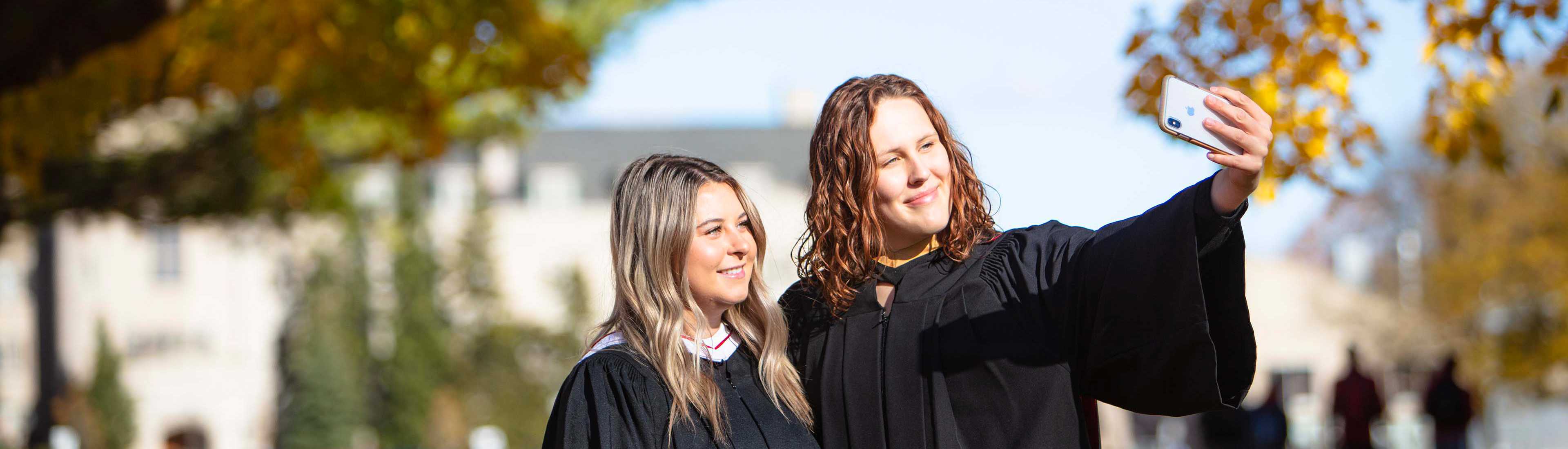 Two university students in graduation gowns taking a selfie on campus during autumn.