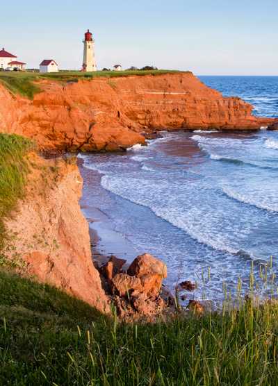 A scenic view of the red cliffs and a lighthouse overlooking the ocean on the Magdalen Islands in Quebec, Canada, with gentle waves crashing against the shore.