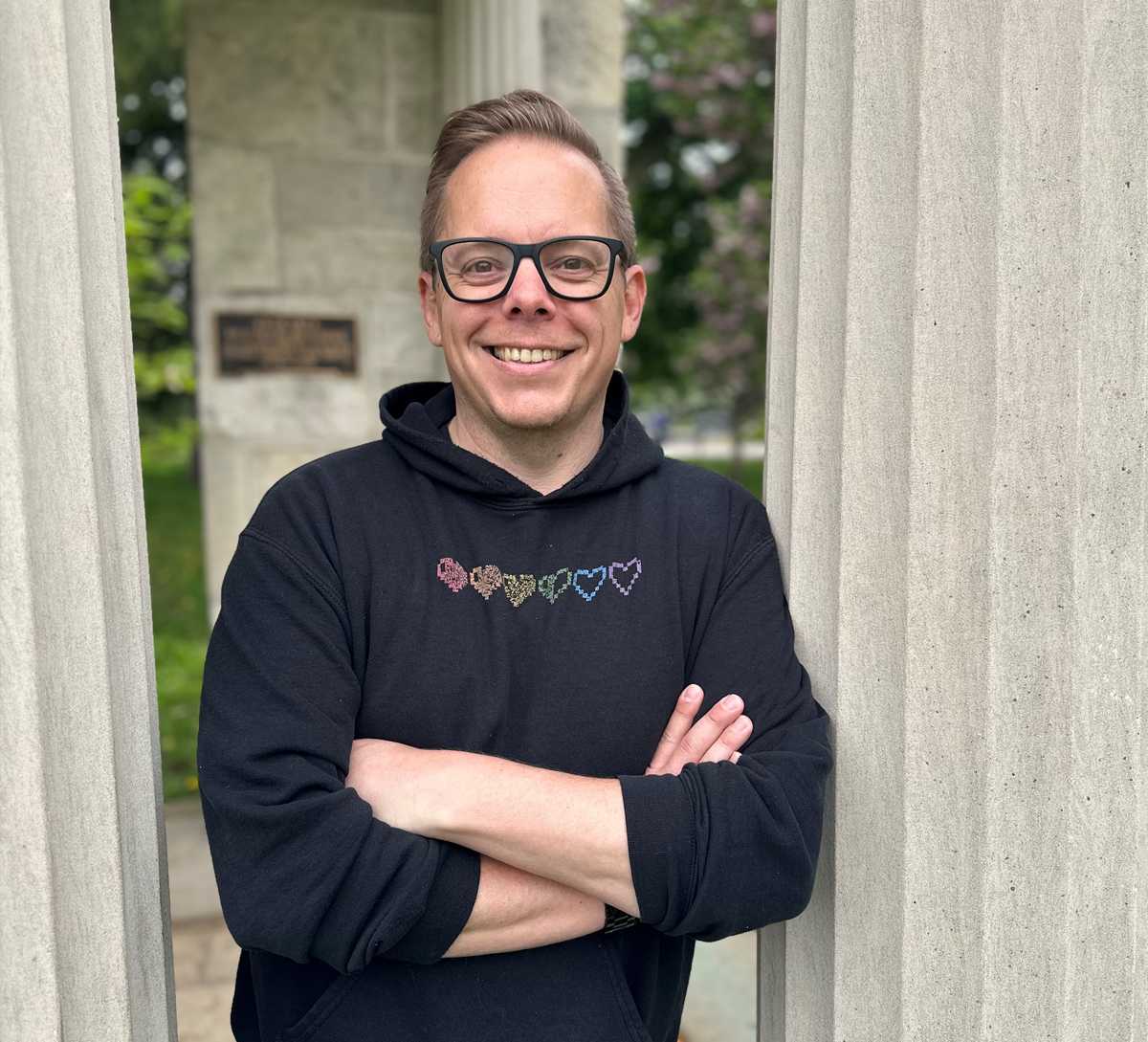 Dan Gillis, wearing glasses and a black hoodie with a colorful heart pattern, standing outdoors with arms crossed between two stone pillars, smiling at the camera.