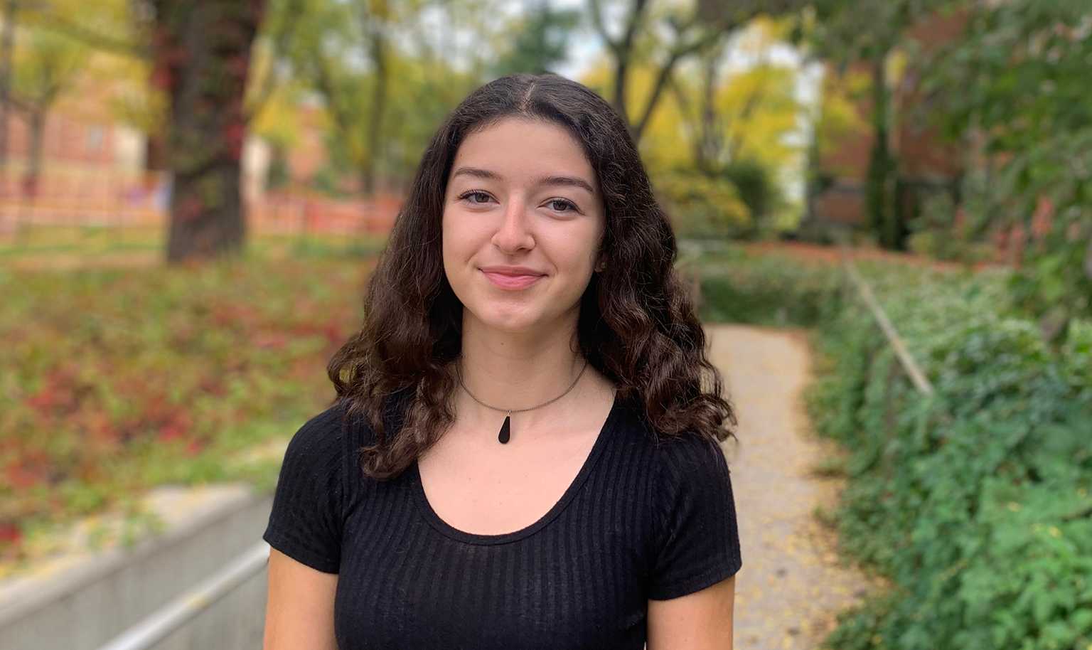 A smiling young woman with curly brown hair standing outdoors on a college campus pathway, surrounded by green foliage and autumn-colored trees. She is wearing a black top and a simple necklace, exuding a friendly and approachable demeanor.