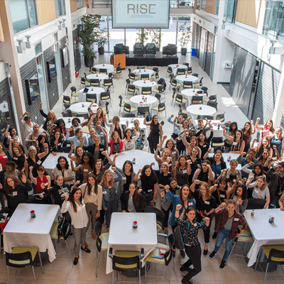 A large group of diverse women at the RISE Conference gathered in a bright, modern atrium, raising their fists in a show of empowerment and solidarity. The event setup includes round tables, creating a welcoming atmosphere for networking and discussion.