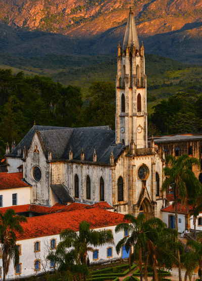 The Santuário do Caraça, a historic Gothic-style church in Brazil, featuring a tall spire, arched windows, and intricate detailing, surrounded by red-roofed colonial buildings and lush palm trees, set against a dramatic mountain landscape at sunset.