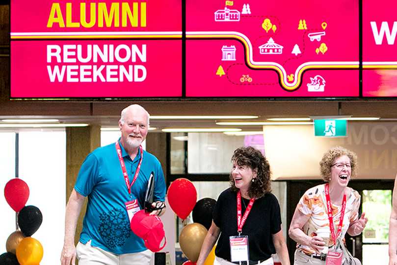 Group of alumni smiling and walking under a bright sign that reads 'Alumni Reunion Weekend,' with festive balloons and colorful event decorations in the background