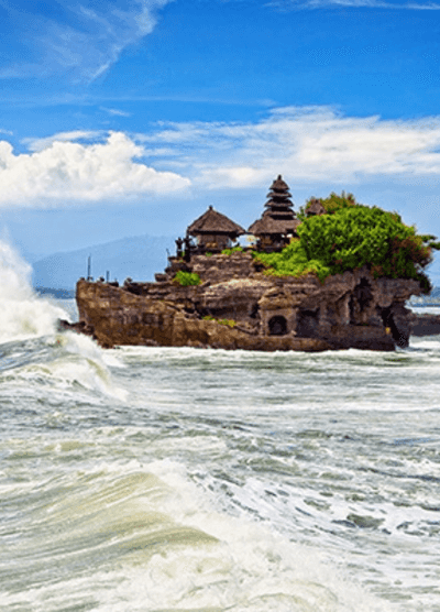 The Tanah Lot Temple in Bali, Indonesia, sit atop a rocky outcrop surrounded by crashing ocean waves. The clear blue sky and lush greenery contrast with the powerful surf, highlighting the temple’s stunning coastal location.