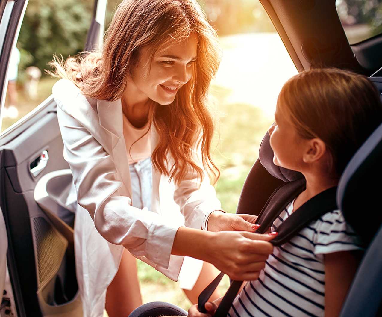 A smiling woman helps a young girl buckle her seatbelt in a car seat, with sunlight streaming through the car door.