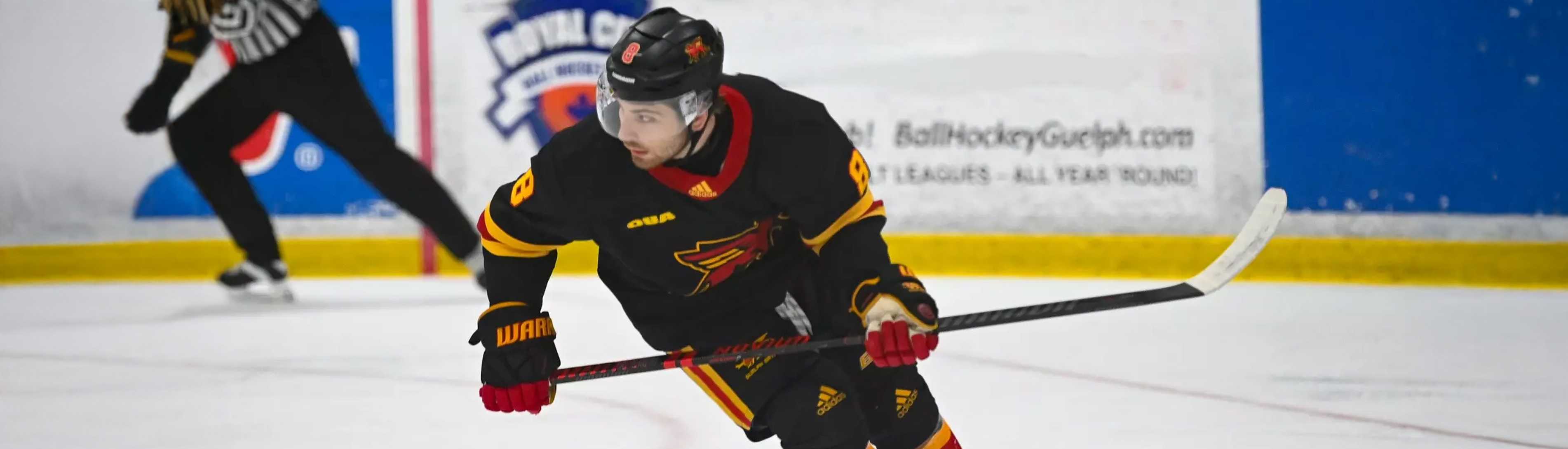 A Guelph Gryphons men’s hockey player in a black and red uniform skates down the ice, focused on the play while holding his stick. A referee is visible in the background near the boards.