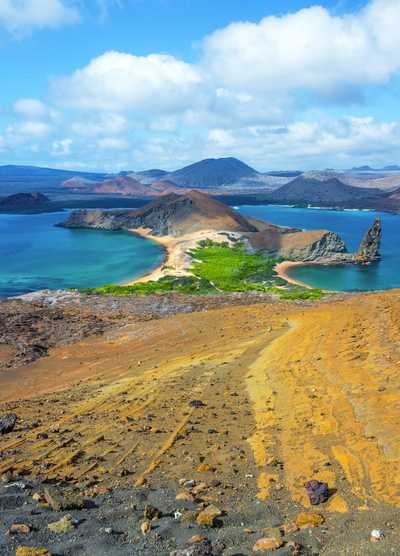 Panoramic view of Bartolomé Island in the Galápagos Islands, featuring a rugged volcanic landscape with golden and reddish terrain, turquoise waters, green vegetation near the shoreline, and the iconic Pinnacle Rock rising from the sea under a bright blue sky with scattered clouds.