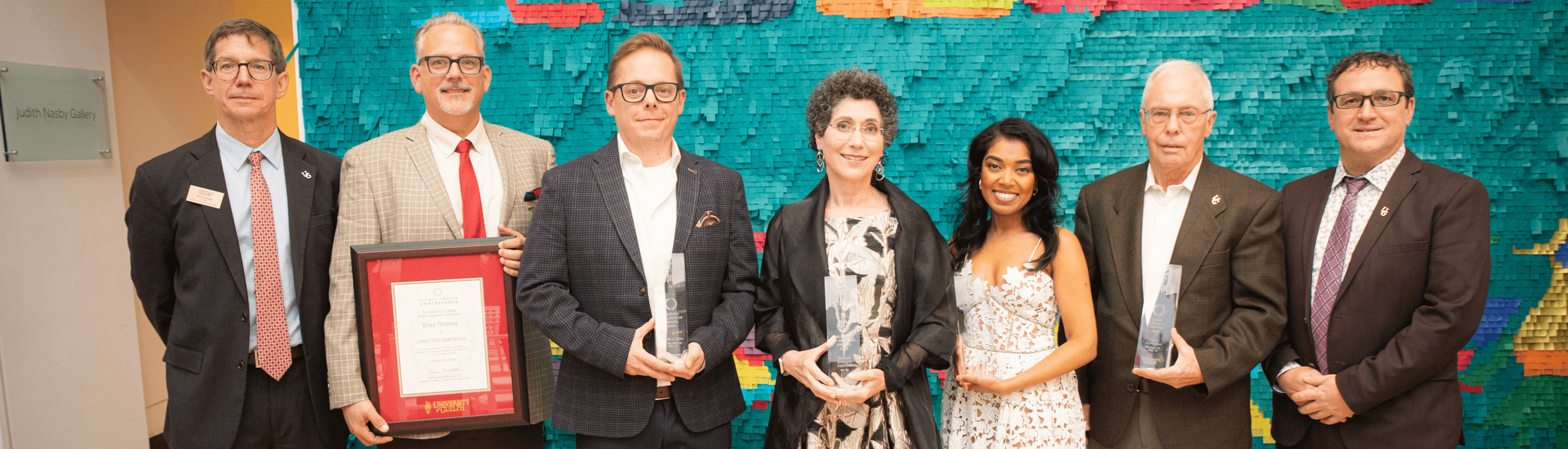 Group photo of award recipients and University of Guelph Alumni Association at the Art Gallery of Guelph. Seven people stand side by side, some holding awards and framed certificates, with a colourful mosaic background