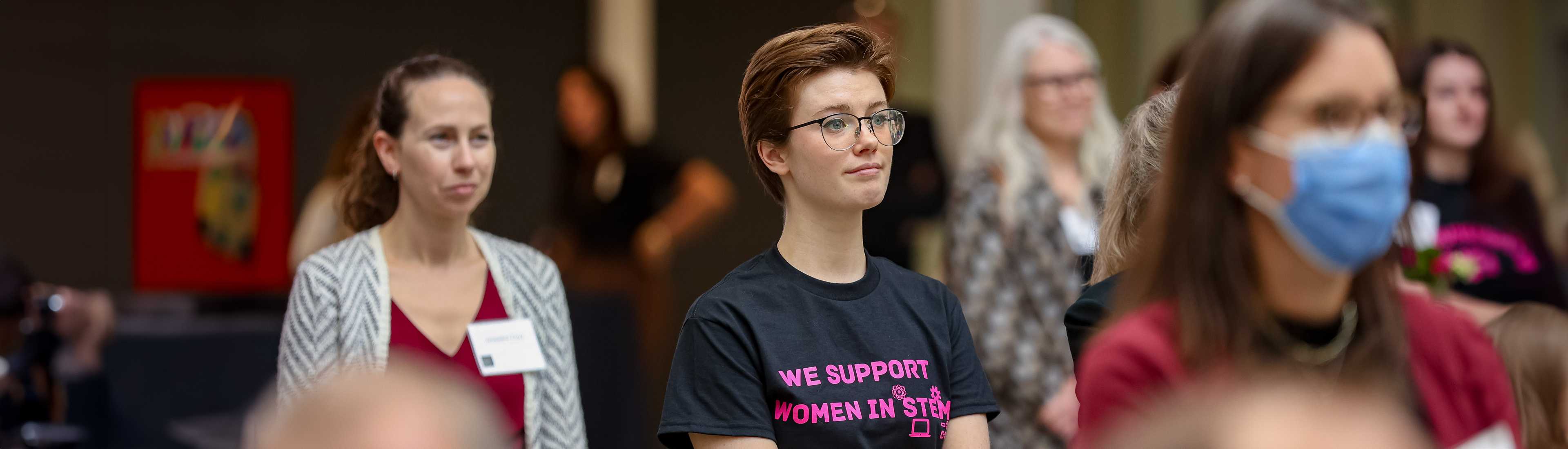 Group of individuals attending a STEM event, with one person wearing a black t-shirt that reads 'We Support Women in STEM' in bold pink text.