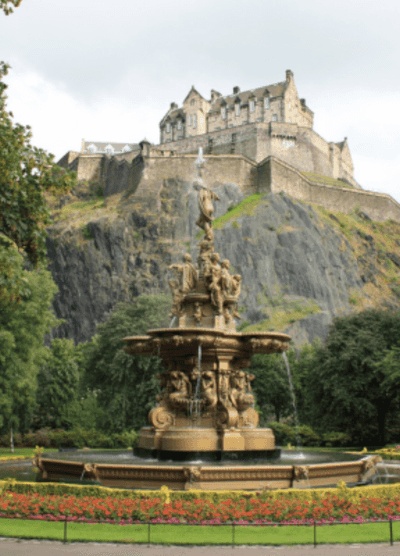 The ornate fountain in Princess Street Gardens, Edinburgh, with the historic Edinburgh Castle perched on the hill behind it. The fountain features intricate sculptures and flowing water, set against a backdrop of lush greenery and the castle’s dramatic stone walls.