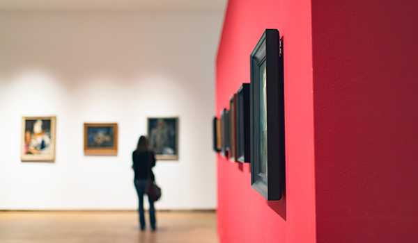 Person standing in an art gallery viewing framed paintings on a white wall, with a red accent wall in the foreground.