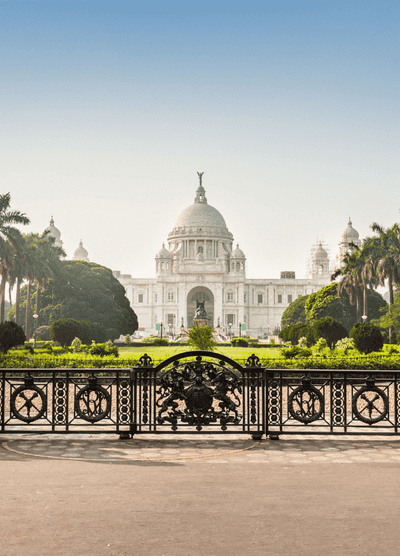 e Victoria Memorial in Kolkata, India, a grand white marble structure with a central dome, surrounded by lush gardens and palm trees, viewed from behind an ornate black wrought-iron gate.